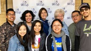 Students and Pastor Jeanne Roe Smith pose in front of a step and repeat that say 501 Cafe.