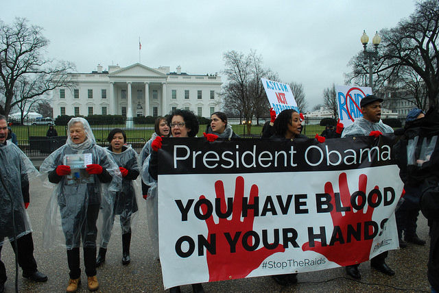 Protesters wearing red gloves hold a sign that reads, "President Obama: You have blood on your hands. #StopTheRaids" outside of the White House.