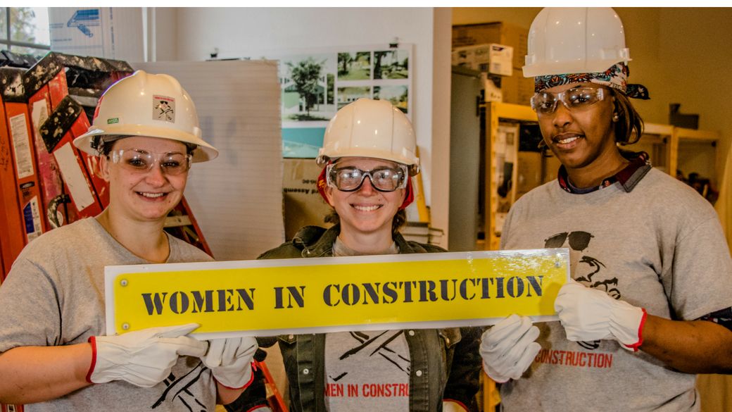 Three women wearing safety equipment hold a yellow sign which reads "women in construction." 