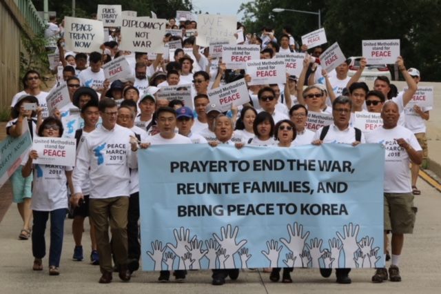 Marchers walk on a street holding signs that read "United Methodists stand for peace," and "Peace treaty now," and "Diplomacy not war." The marchers in front of the group carry a blue banner that reads, "Prayer to end the war, reunite families, and bring peace to Korea." 