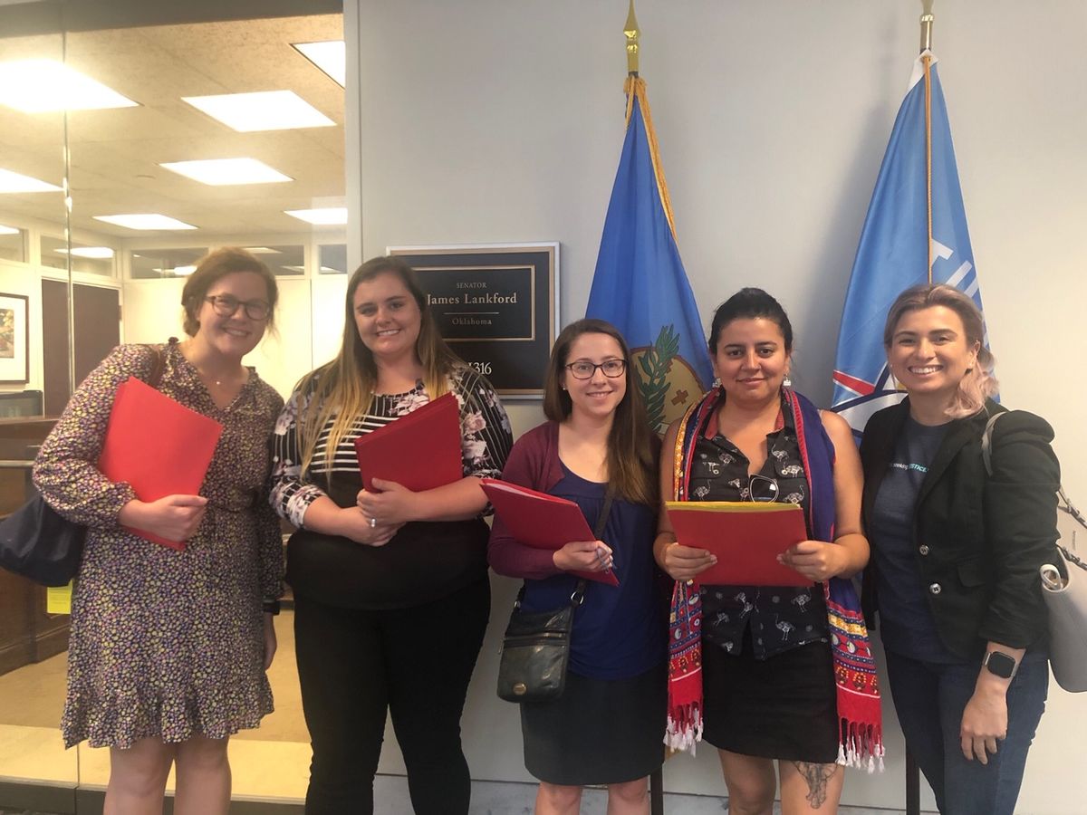 Five people stand in front of an office door. Four of them are holding folders. Behind them are two flags and a plaque that reads, "Senator James Lankford, Oklahoma." 