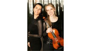 The Carr-Petrova Duo, both wearing black, pose. One has brown hair the other blond. The blond-haired woman holds a violin. 