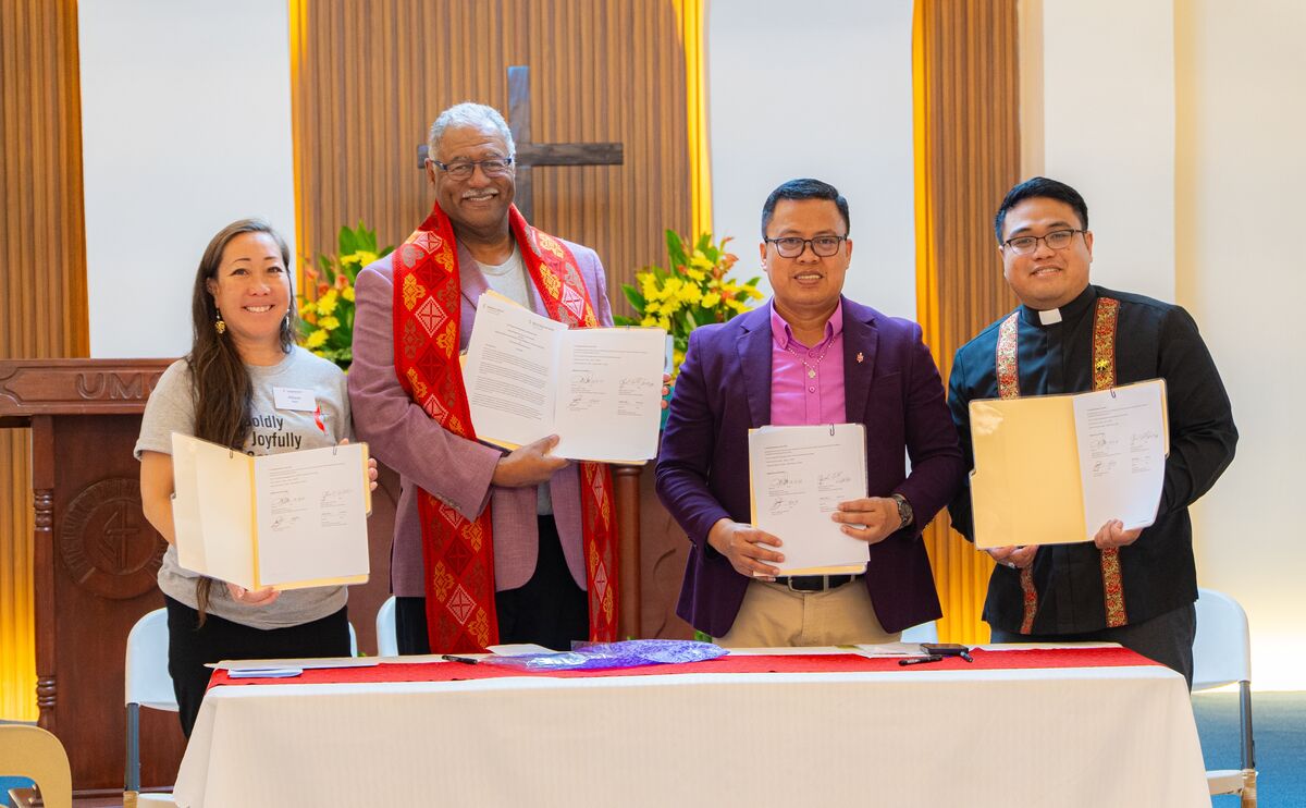 Four people stand in a row behind a long table, each holding up a copy of an agreement. In the background is a crucifix affixed to the wall.