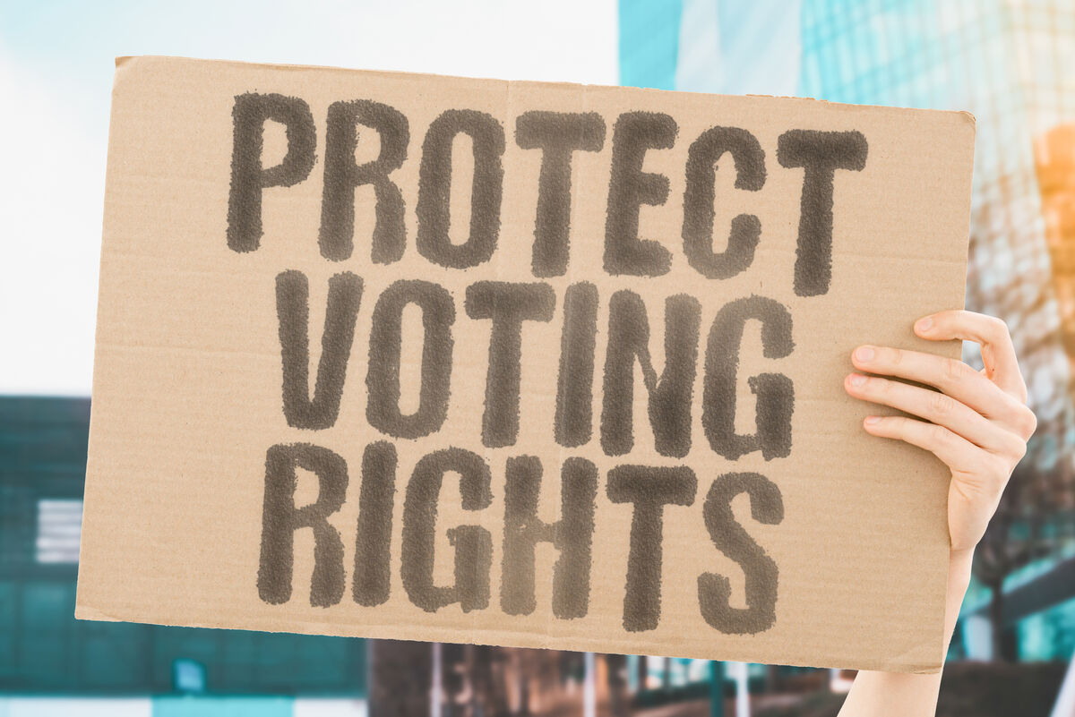 A hand holding up the sign "Protecting Voting Rights," written on cardboard in inky letters.