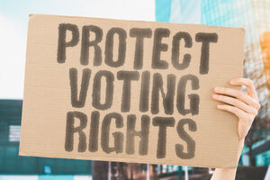 A hand holding up the sign "Protecting Voting Rights," written on cardboard in inky letters.