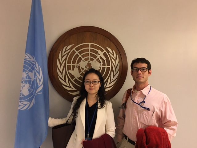 Two fellows stand in front of the U.N. flag and symbol. 